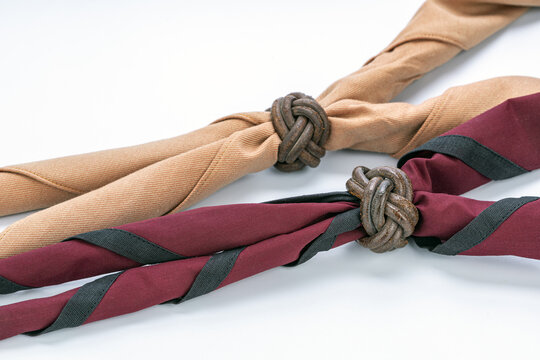 Traditional Thai boy scout neckerchiefs in khaki tan and maroon with black stripe fastened by handcrafted braided leather Turk's head woggle knots displayed on clean white studio background