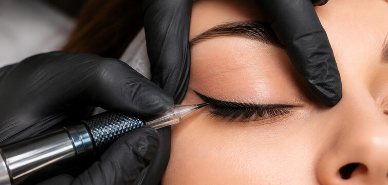 Cosmetologist performing permanent eyeliner tattoo on woman eyelid. Professional tattooist using electric machine for micropigmentation on client eye. Beauty salon permanent makeup artist.