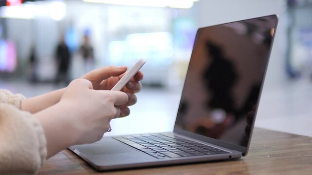 Person holds phone near laptop. Hands interact with devices. Background blurred in public space. Focus on multitasking with tech tools. Wood table surface under devices