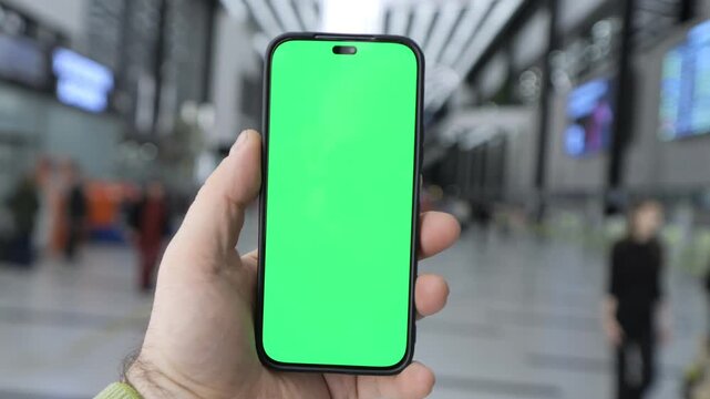 Hand holds smartphone with green screen. Background shows busy train station. Blurred people walk around. Phone screen remains green throughout. Urban setting with large digital displays