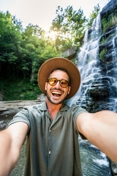 Handsome tourist taking selfie picture in front of waterfall - Happy hiker exploring forest - Traveling life style concept with happy man wearing hat and sunglasses enjoying freedom in the nature 