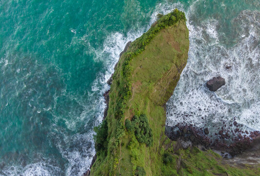 Aerial view of a lush green coastal cliff with a narrow path, surrounded by turquoise ocean waves crashing against the rocky shoreline in Kebumen, Central Java, Indonesia.