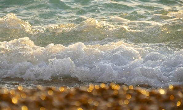 Big sea wave splash with foam on golden sparkle beach with pebble in sunset - summer sea background