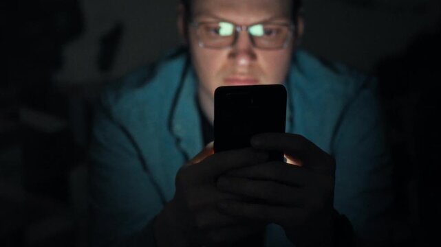 A man with glasses holds a phone, staring at its bright screen in the dark night. His scrolling reveals a harmful smartphone dependence, showing how mobile devices can disrupt healthy sleep patterns
