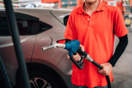 close-up, selective-focus shot at a gas station. An attendant, wearing a bright orange polo shirt and a black arm sleeve, holds out a fuel pump nozzle with a blue handle