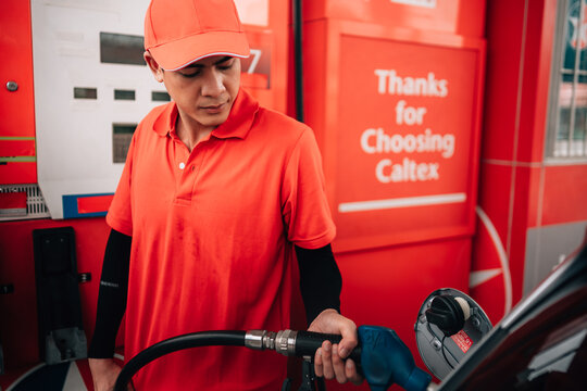 a male gas station attendant in the process of refueling a vehicle.He is looking down attentively as he holds a blue-handled fuel nozzle inserted into the open fuel tank of a dark-colored car.