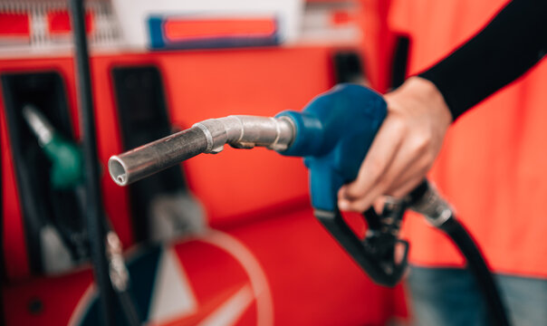 close-up, selective-focus shot at a gas station. An attendant, wearing a bright orange polo shirt and a black arm sleeve, holds out a fuel pump nozzle with a blue handle
