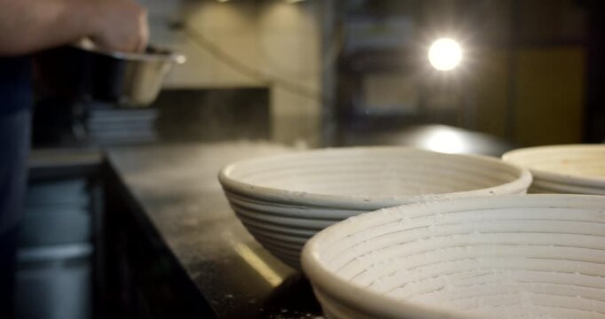 Baker sprinkling flour over proofing baskets in a commercial kitchen countertop