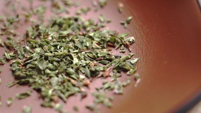 Aromatic crushed dried oregano leaves cascade into a rustic brown ceramic bowl in slow motion. Green organic herb flakes. Classic Mediterranean seasoning. Rotation