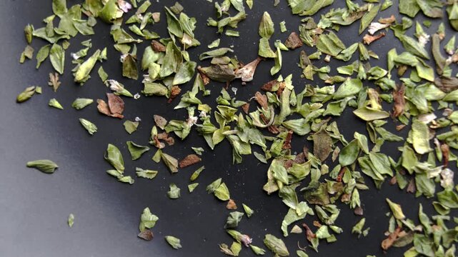 Scattered dried oregano (Origanum vulgare) leaves on a black surface. Mediterranean seasoning herb. Rotation. Top view