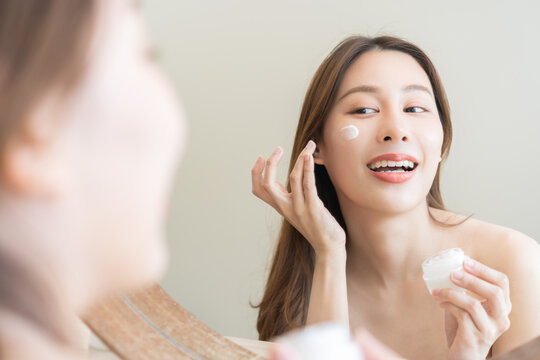 Facial beauty skin care, smile of pretty asian young woman in bathrobe looking at mirror, hand applying moisturizer lotion on her face, holding jar of skin cream before makeup cosmetic routine at home