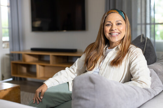 Mature African American woman sitting on gray sofa in living room, smiling, flat-screen TV visible