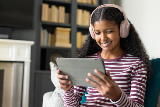 Teenage African American female sitting on couch using tablet with pink headphones at home