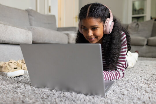 African American teenage female lying on shag rug, using silver laptop and wearing pink headphones