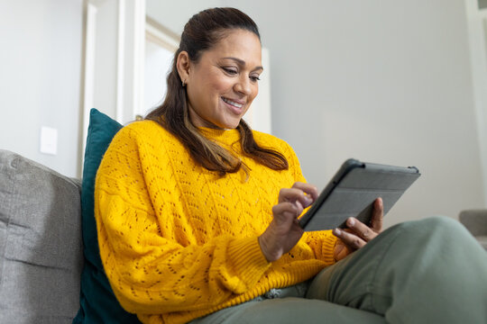 African woman sitting on gray sofa with teal pillow wearing mustard knit sweater tapping tablet