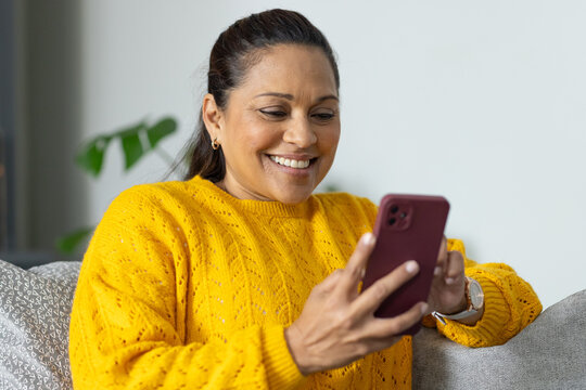 African American woman sitting on gray sofa in yellow sweater, holding burgundy phone