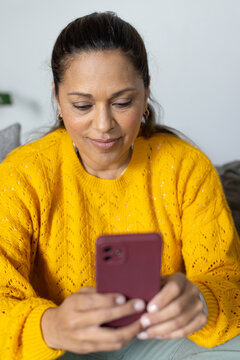 African woman scrolling maroon smartphone while sitting on living room sofa in yellow knit sweater