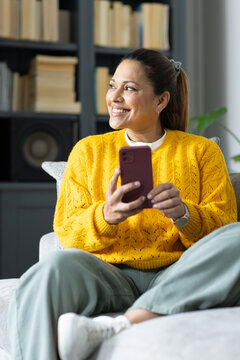 African American woman in 40s sitting on light couch yellow-sweater holding maroon-phone, bookshelf