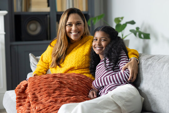 Mother and daughter sitting at home on gray sofa sharing orange throw wearing mustard sweater