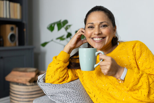 Mature African woman reclining on sofa in living room holding mint mug, smiling in mustard sweater