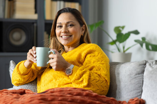 Mature African American woman smiling while holding green mug on gray sofa wearing yellow sweater