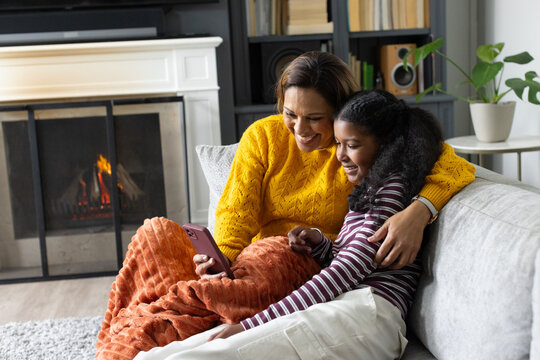 African American mom and teenage daughter holding phone with pink case on sofa with orange throw