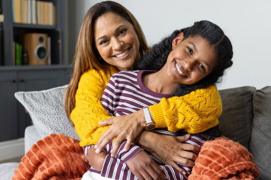 African American mother hugging teenage girl on gray sofa at home wearing yellow sweater with books