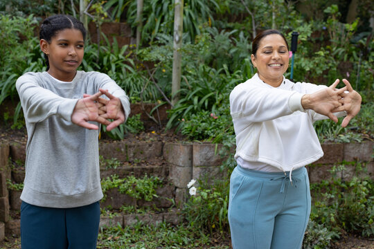 African American mother and teenage daughter stretching arms in garden by stone wall, in activewear