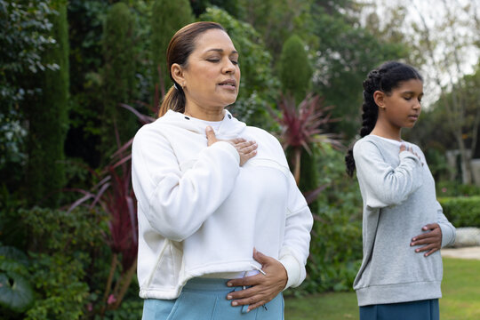 African American mother and daughter practicing breathing exercises in garden with trimmed hedges
