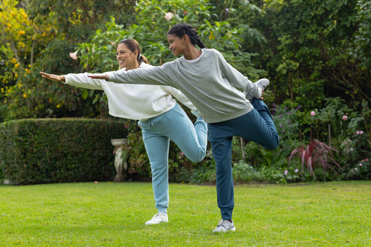 African mother and teenage daughter stretching on garden lawn wearing sweats and sneakers by statue