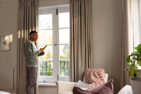 African American man standing left of French doors, holding smartphone and mug, copy space