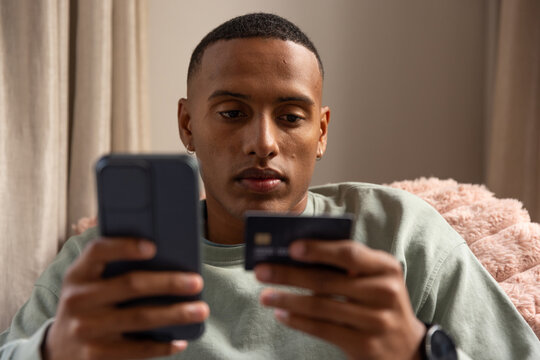 African man sitting in plush armchair, holding smartphone and card, wearing pale green sweatshirt