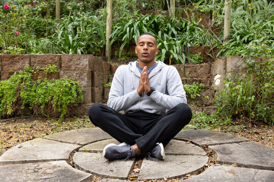 African American man sitting crosslegged on stone patio meditating, pressing palms, wearing hoodie
