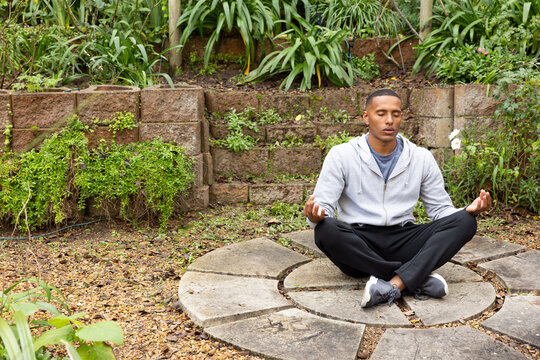 African male meditating on stone circle in backyard wearing hoodie, sneakers, copy space