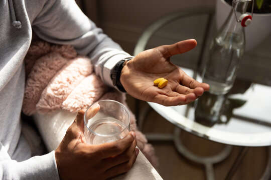 Male in hoodie sitting on armchair holding glass of water showing two yellow pills home