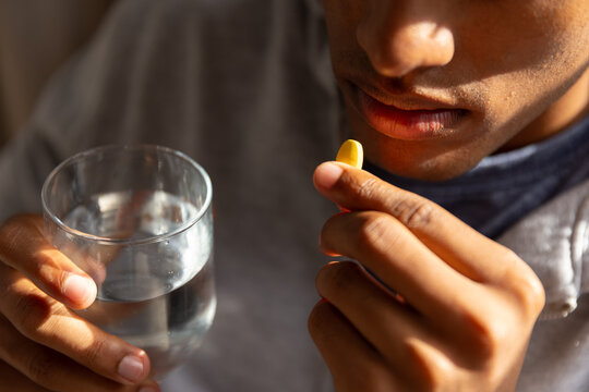 African American man holding yellow capsule near lips and water glass at home wearing gray shirt