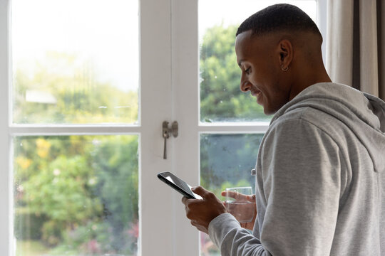 Man in light gray hoodie standing by glass doors holding smartphone, drinking glass, copy space