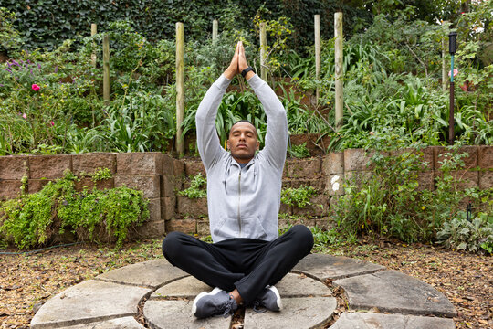 African-American man sitting cross-legged on stone-platform in garden, wearing hoodie lifting hands