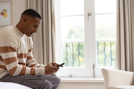 African American man sitting on bed edge holding smartphone and wearing striped sweater in bedroom