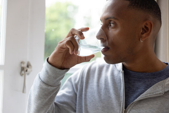 Man wearing gray hoodie standing by white-framed window with latch, holding glass