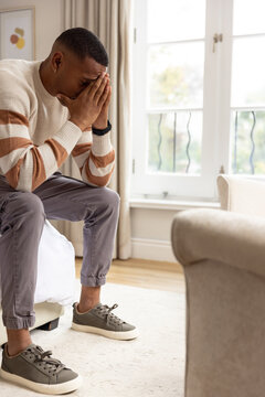 African man in 20s sitting on bed edge bowing head covering face wearing wristwatch and sneakers