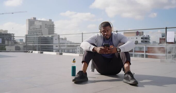 Mid-adult African man in hoodie postrun adjusting headset sipping bottle checking phone on rooftop