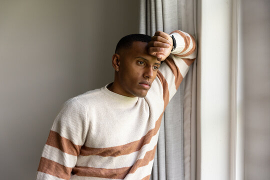 African American man leaning on window at home, wearing striped sweater and watch, gazing outward