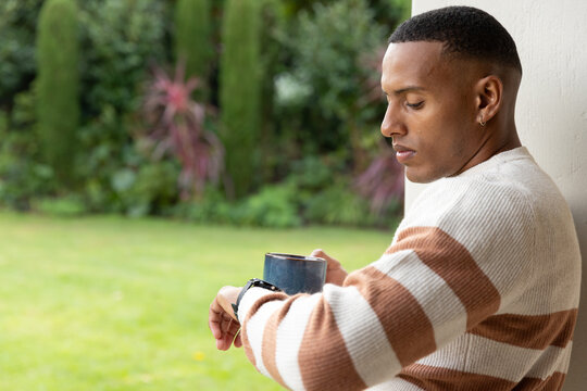 Man 20s leaning at porch wall holding dark mug checking watch wearing striped sweater, copy space