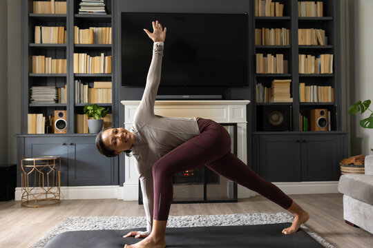 Adult woman of African descent practicing deep lunge on yoga mat in living room, smiling