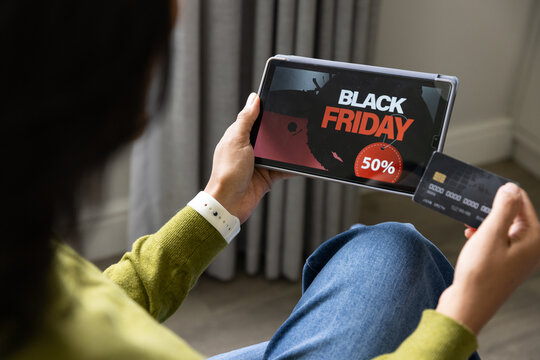 African woman wearing white watch, holding tablet showing BLACK FRIDAY 50% and using card at home