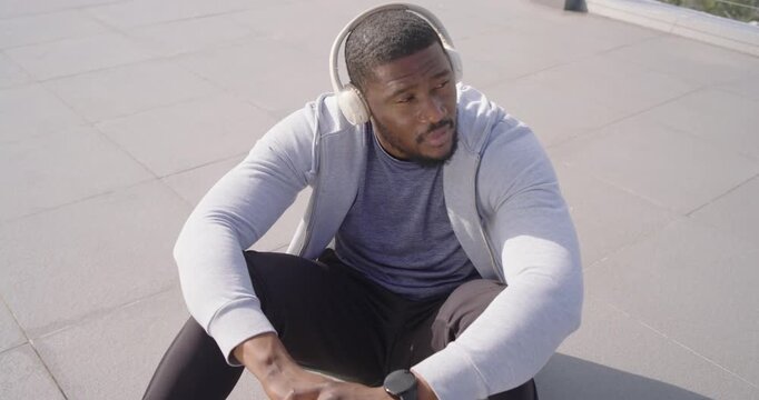 Mid-adult African man in hoodie with headphones checking smartwatch after scanning rooftop horizon