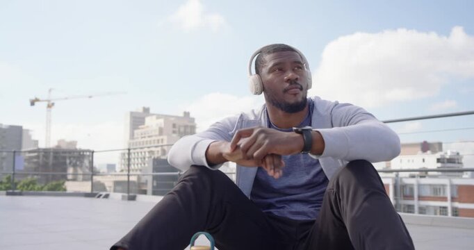 Mid-adult man sitting on rooftop putting on headphones, listening to music and scrolling smartphone