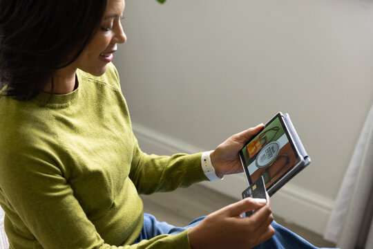 Adult African American woman wearing green sweater holding tablet, credit card at home, copy space