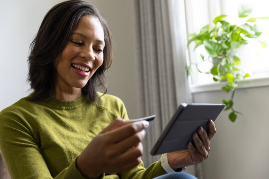 Adult African American woman sitting at home, wearing green sweater, using tablet and credit card